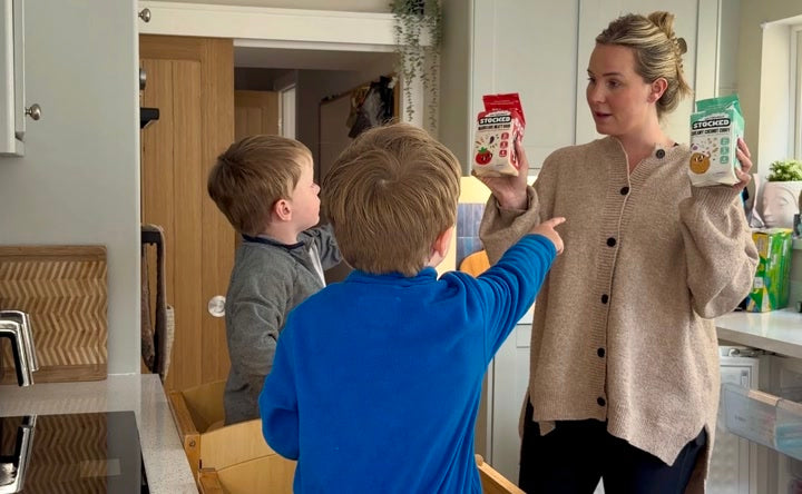 A mum holding up two STOCKED packs while her two young boys reach for them in the kitchen