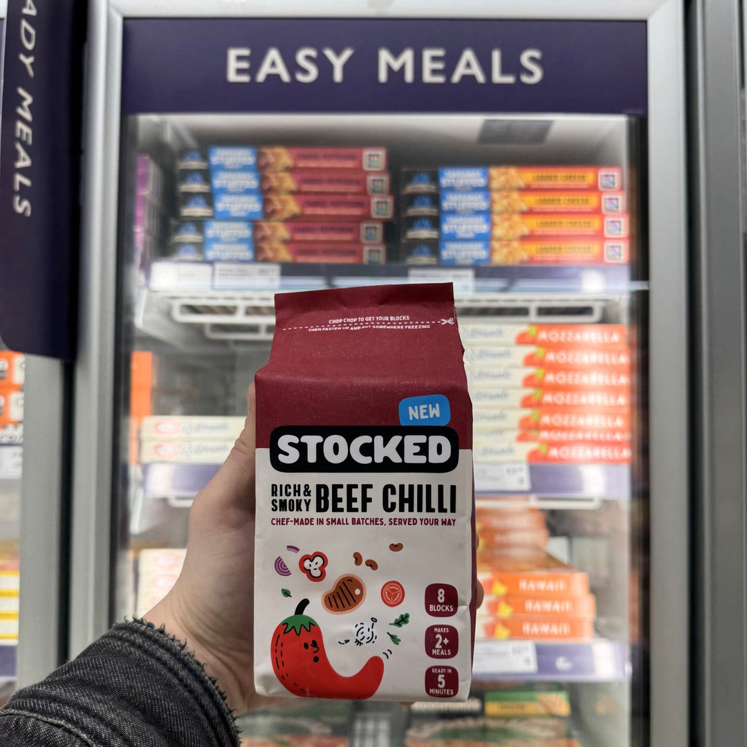 A Stocked Beef Chilli pack held up in front of a supermarket freezer of ready meals